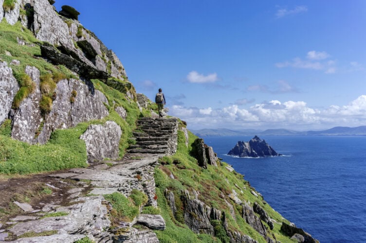A person walks up stone steps on a rocky cliffside overlooking the ocean with distant islands under a blue sky.