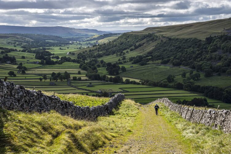A person walks along a gravel path flanked by stone walls, overlooking green fields and rolling hills typical of England countryside regions under a cloudy sky.
