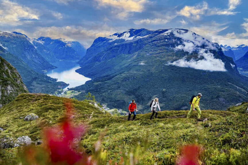 Three hikers walk uphill on grassy terrain in Norway, with mountains, a lake, and a cloudy sky in the background. Red flowers are visible in the foreground, highlighting why this is the best time to visit.