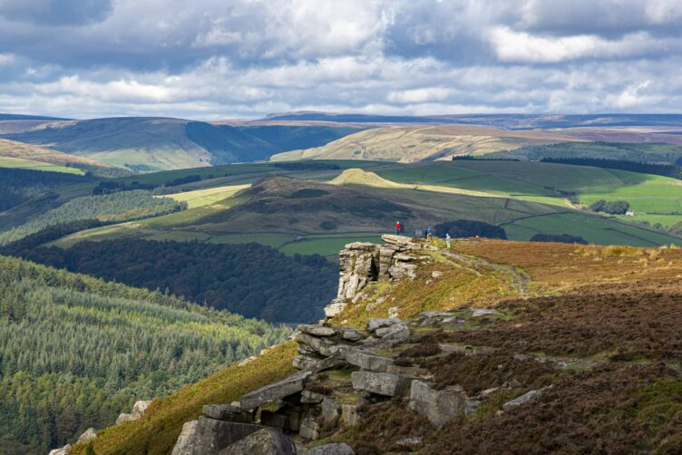 People stand on a rocky outcrop overlooking rolling green hills and patchwork fields, soaking in the beauty of England’s countryside regions beneath a partly cloudy sky.
