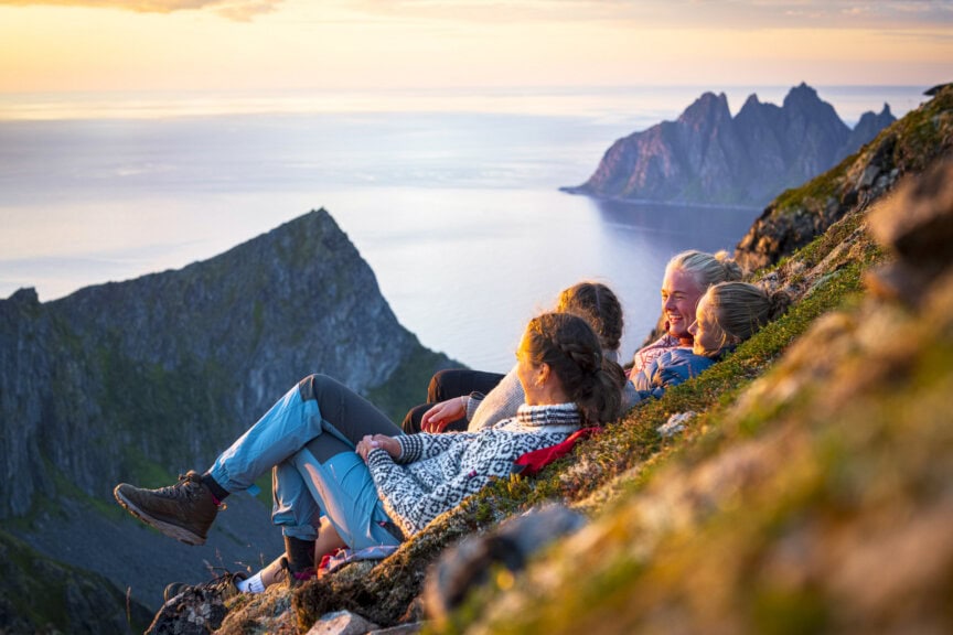 Four people sit on a grassy mountainside in Norway, overlooking the sea and distant peaks at sunset, appearing relaxed and enjoying the view—truly capturing the best time to visit this breathtaking destination.