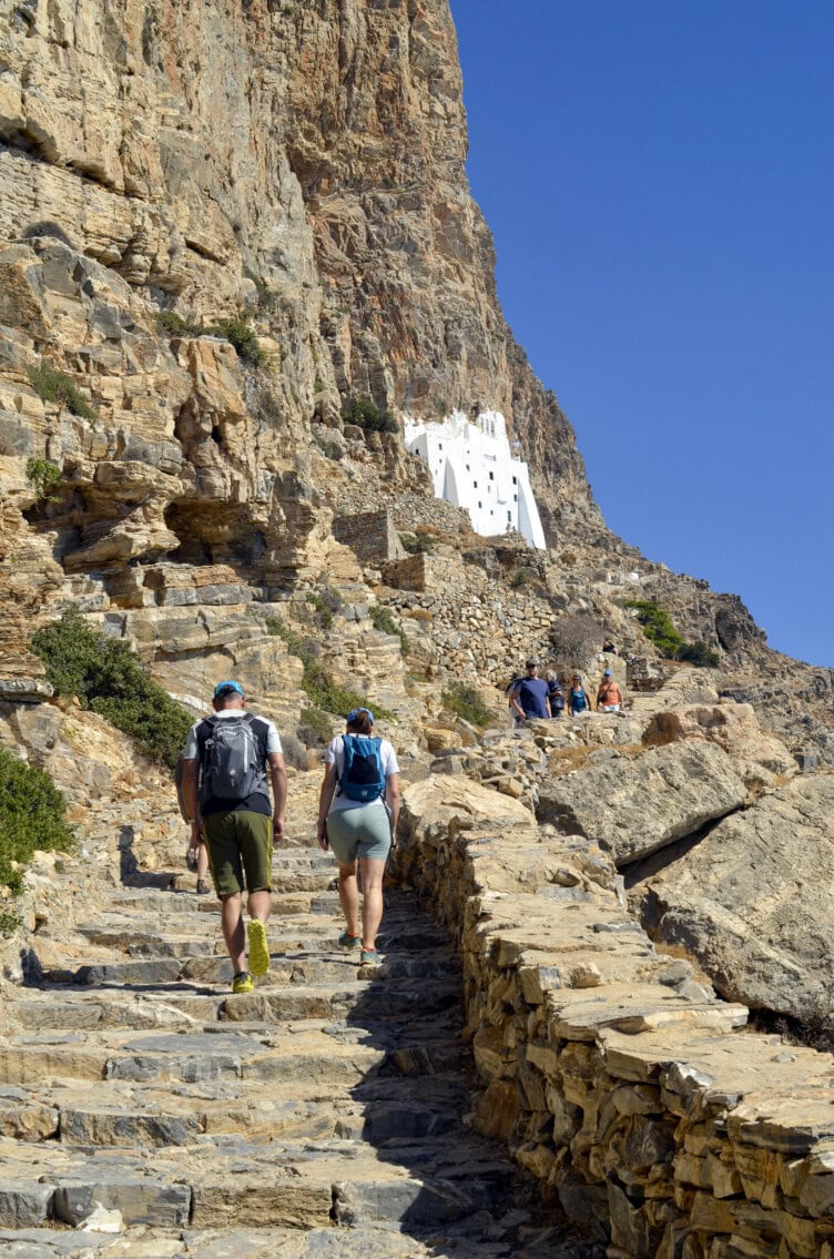 Hikers ascend a stone staircase along a rocky cliff toward a white monastery built into the mountainside under a clear blue sky.
