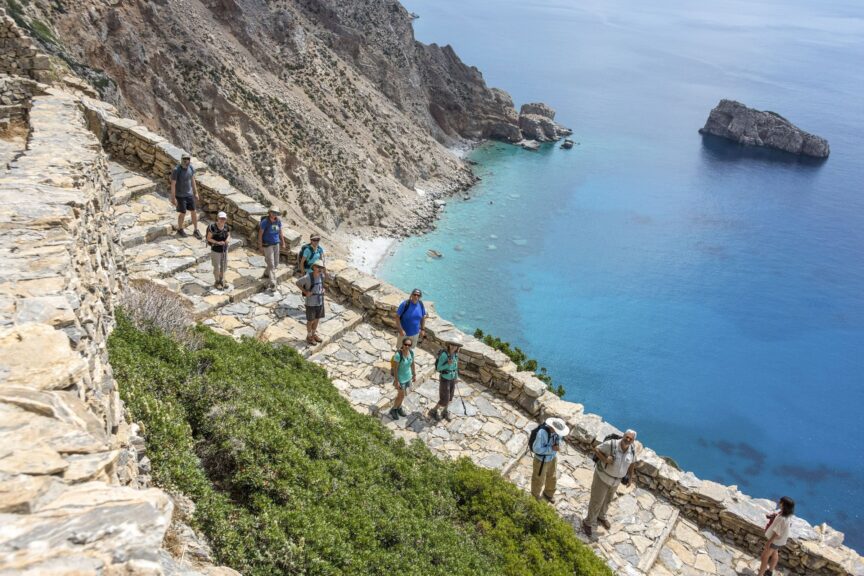 Hikers walk along a stone path on a steep cliff above clear blue water, with rocky coastline and shrubbery visible—the perfect scene to experience during the best time to visit Greece.
