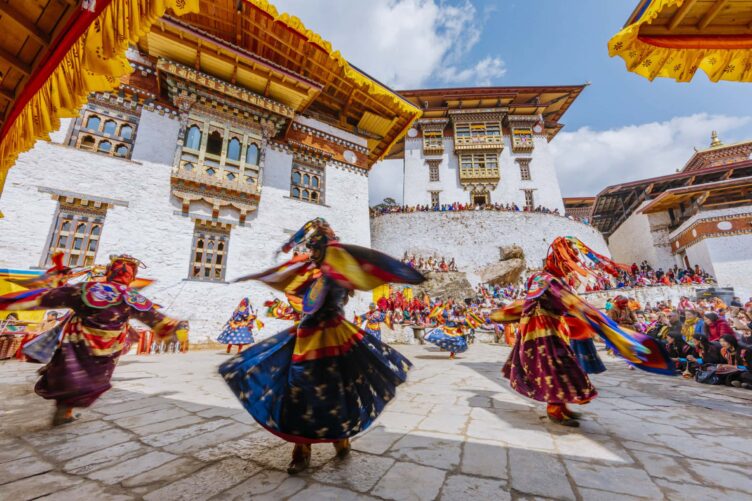 Dancers in colorful traditional costumes perform in a courtyard surrounded by Bhutanese architecture, with an audience watching under a bright sky.