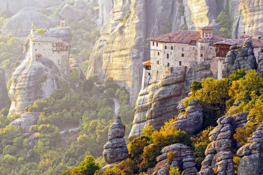 Two historic monasteries stand atop tall, rocky cliffs surrounded by greenery in Meteora, Greece, under soft sunlight—the best time to visit Greece for breathtaking views and tranquil moments.