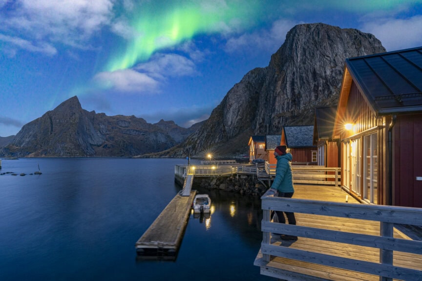 Person standing on a wooden deck by water in Norway, looking at the Northern Lights above mountains and red cabins during twilight—a perfect scene to experience if you’re wondering about the Best Time To Visit this magical destination.