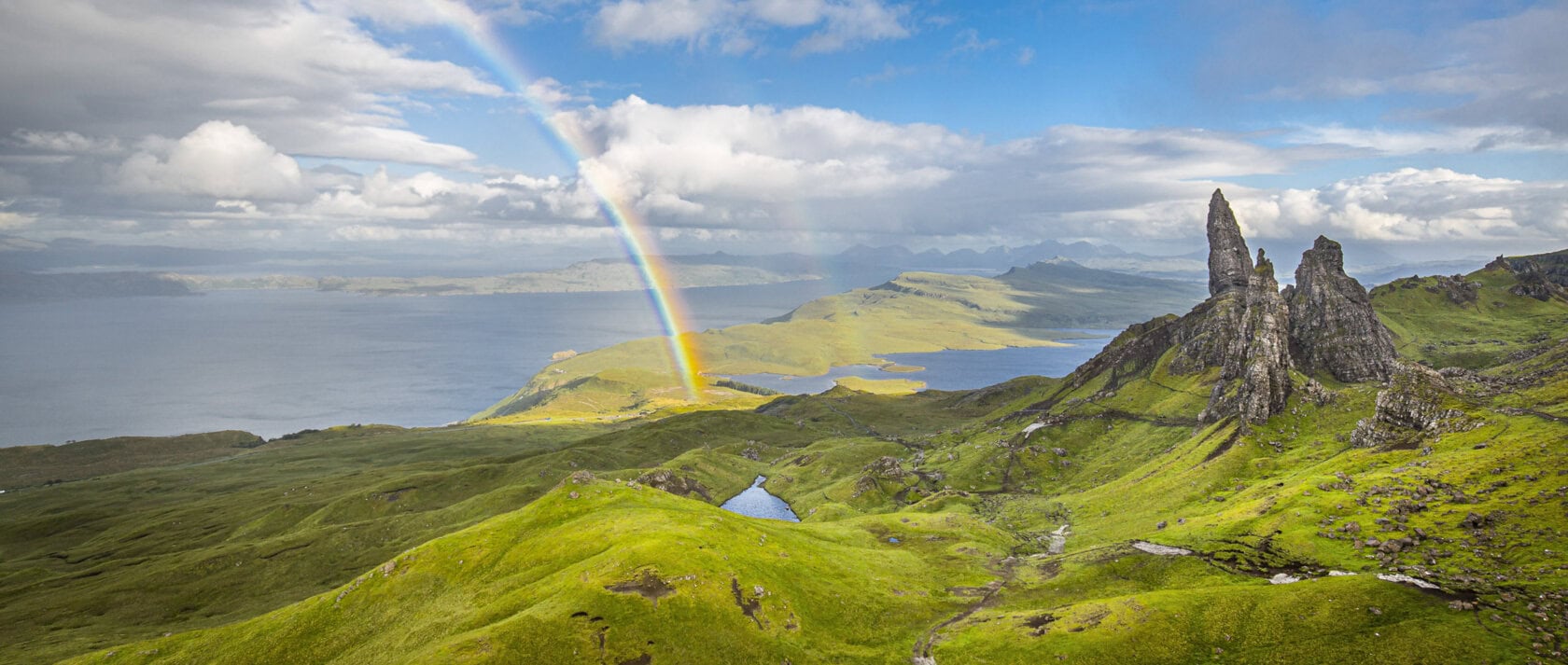 A rainbow arches over lush green hills and rocky formations near the coastline under a partly cloudy sky, showcasing the stunning landscapes that await during the best time to visit Scotland.