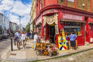 People browsing items outside Alice’s antique shop on a sunny London street corner; colorful shopfront displays vintage goods and artwork—one of the charming things to do in England.