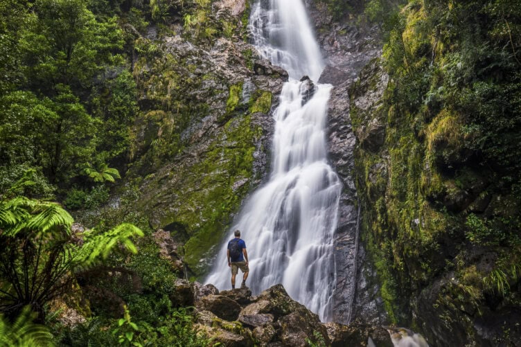 A person with a backpack stands on rocks at the base of a tall, cascading waterfall surrounded by lush green vegetation.