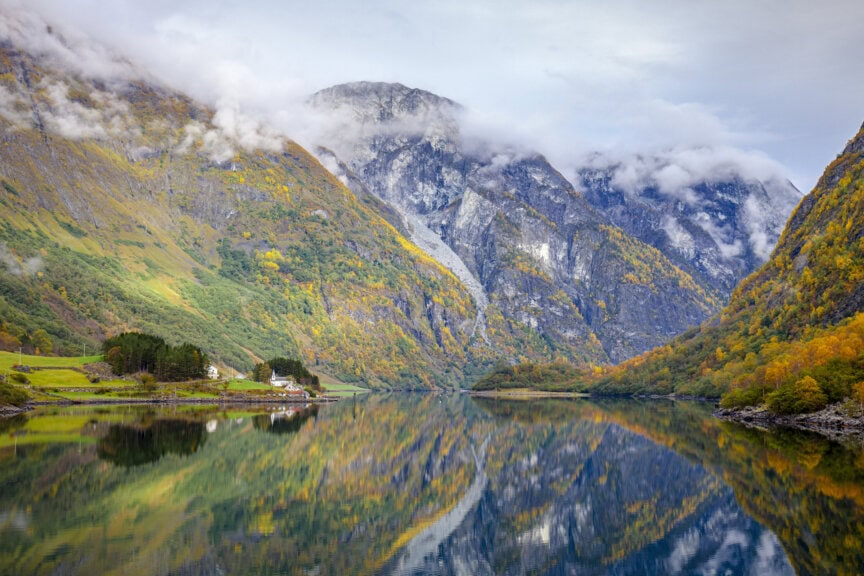 A calm lake in Norway reflects surrounding mountains with patches of autumn foliage, clouds, and a small cluster of houses on the green shoreline—showcasing the best time to visit this serene landscape.