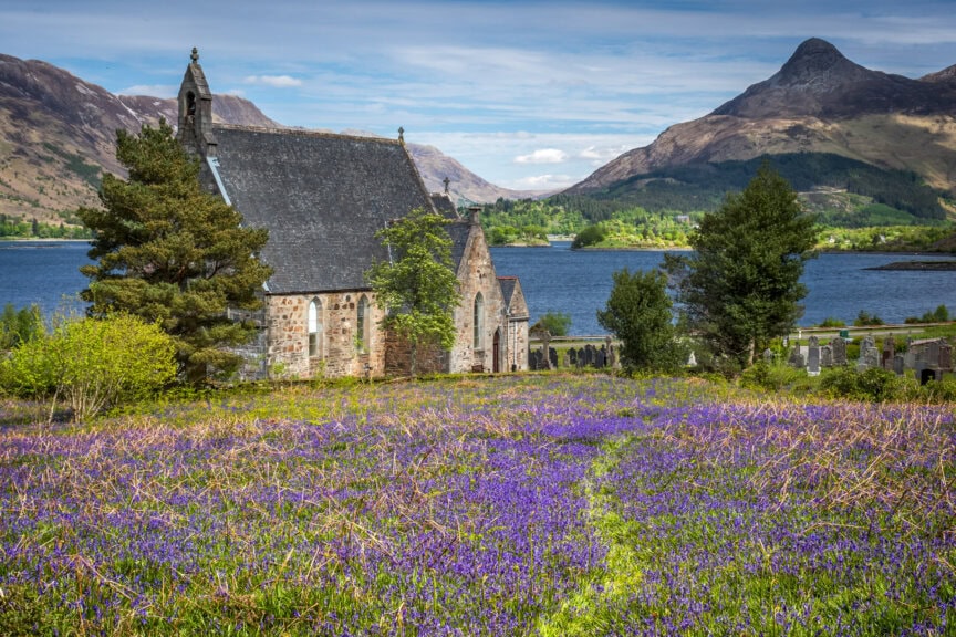 A stone church stands by a lake in Scotland, with mountains in the background, surrounded by trees and a field of purple wildflowers under a partly cloudy sky—the best time to visit is when the flowers are in bloom.
