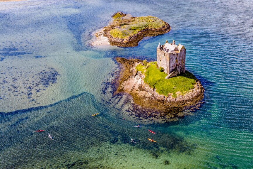 Aerial view of a stone castle on a small island in Scotland, surrounded by clear water, with several kayakers paddling nearby—truly picturesque no matter the best time to visit.