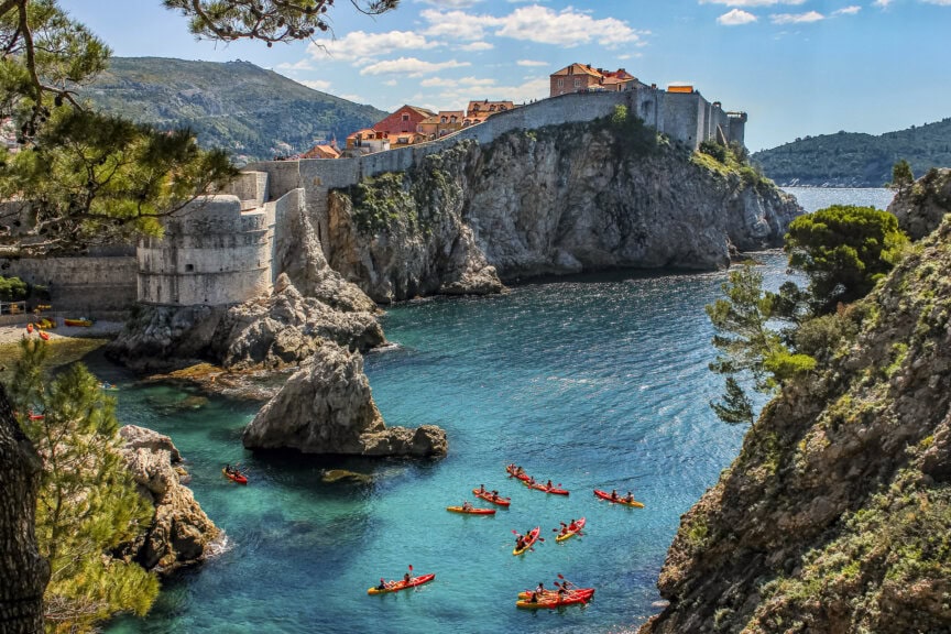 A group of kayakers paddles in clear blue water near rocky cliffs and historic stone buildings on the coast of Croatia under a partly cloudy sky—the best time to visit is during the warm, sunny months.
