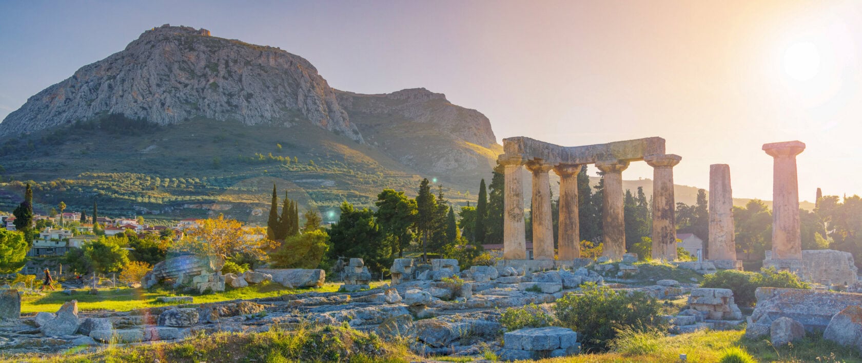 Ancient stone columns of the Temple of Apollo stand in ruins, with mountains and trees in the background under a bright sun—the scene captures why many say it's the best time to visit Greece.