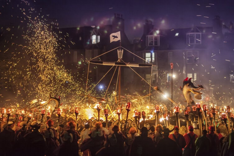 A crowd with torches surrounds a Viking longship set on fire during a nighttime festival in Scotland, sparks flying and buildings aglow—an unforgettable sight and truly the best time to visit for such dramatic celebrations.