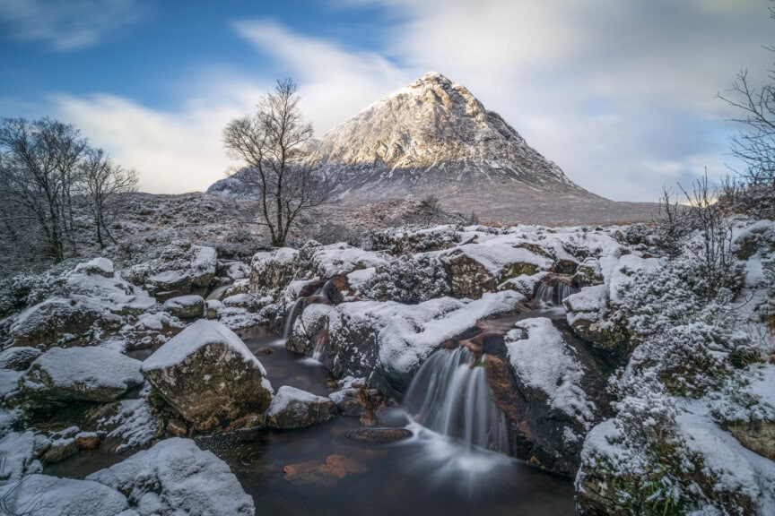 A snow-covered mountain rises behind a rocky stream and leafless trees under a partly cloudy sky in Scotland, offering breathtaking views—especially during the best time to visit.