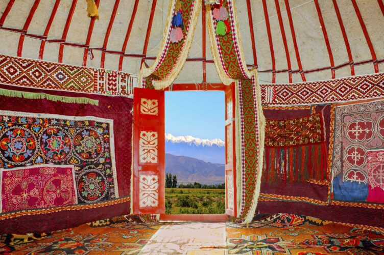 Interior of a colorful yurt with patterned textiles, open door framing a view of green fields and snow-capped mountains under a blue sky.