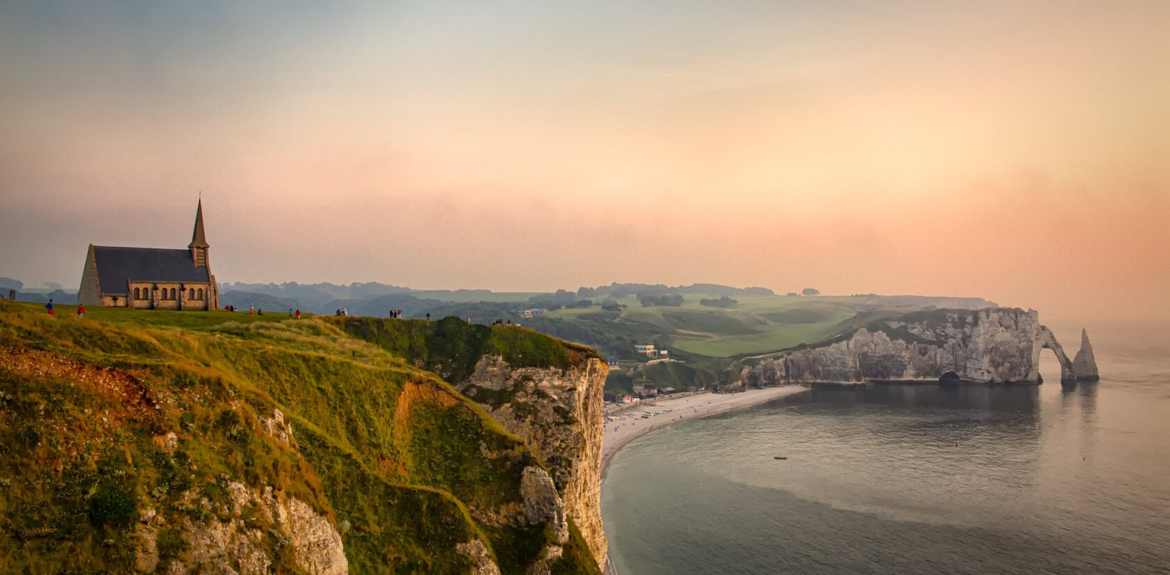 A small church stands on a grassy cliff in France, overlooking a curved coastline and sea at sunset, with rocky formations visible in the distance. Best time to visit is during golden hour for stunning views.