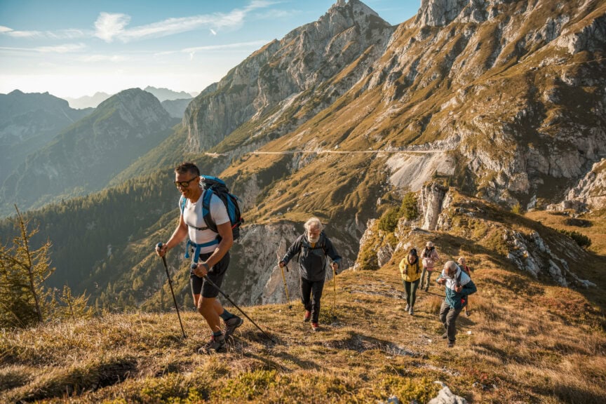 Four people with backpacks hike up a grassy mountain slope using trekking poles, enjoying Slovenia’s rugged mountains and a winding road in the background under a clear sky—the perfect scene for discovering the Best Time to Visit Slovenia.