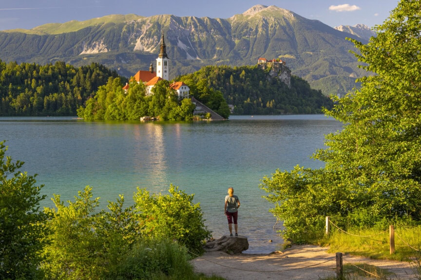 A person stands at the edge of a lake facing an island with a church, surrounded by green trees and mountains—a scene that captures the essence of the Best Time to Visit Slovenia.