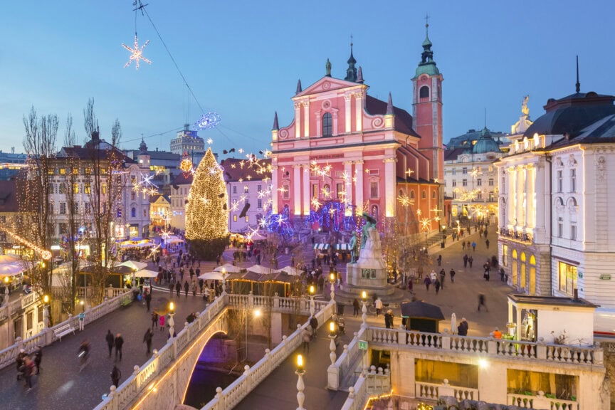 A festive city square with a large decorated Christmas tree, illuminated buildings, and people walking near a pink church at dusk showcases why winter is the Best Time to Visit Slovenia.