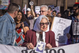 An elderly woman wearing sunglasses and a white headscarf holds a photo at a protest in Buenos Aires, surrounded by others with signs and banners—a powerful glimpse into Argentina's vibrant activist culture.