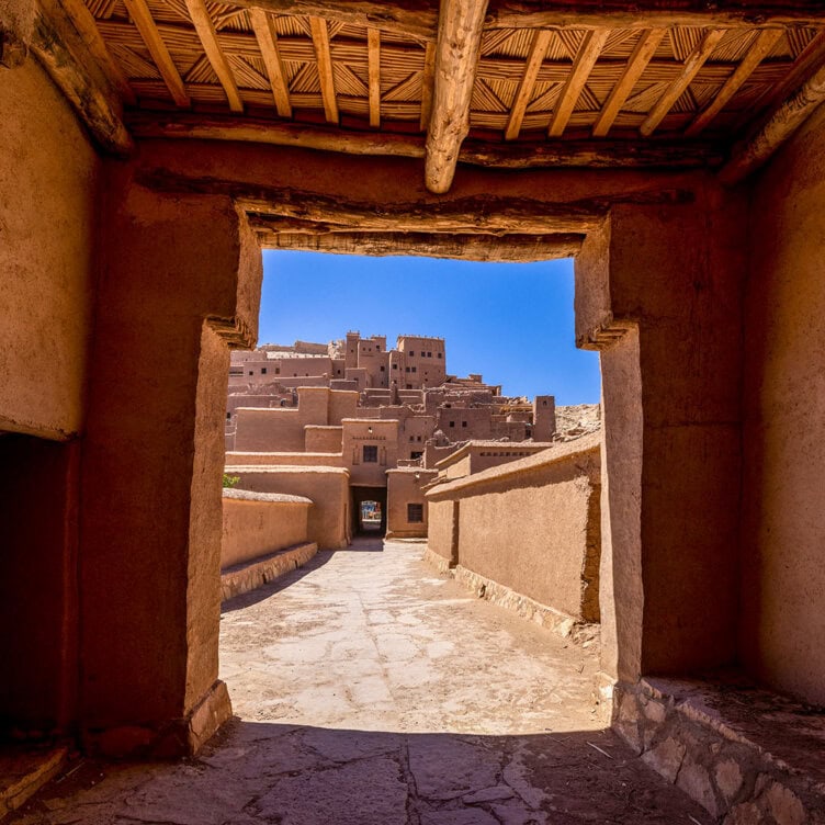 View through an adobe gate leading to a sunlit pathway with traditional mud-brick buildings and blue sky in the background—a glimpse of what to expect when you visit Ait Ben Haddou.