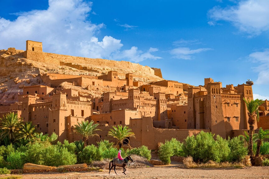 A person riding a horse passes in front of Ait Ben Haddou, an ancient fortified village of brown adobe buildings surrounded by greenery, with a castle-like structure on the hill—just what to expect when you visit this historic site.
