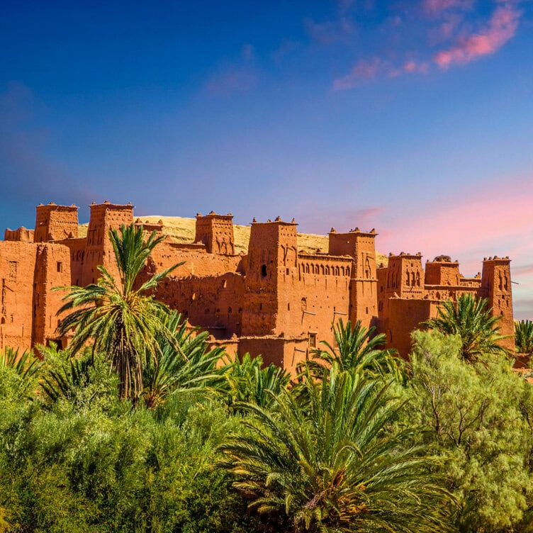 A large, traditional clay fortress with multiple towers rises behind lush green palm trees under a blue and pink sky—just what to expect when you visit Ait Ben Haddou.