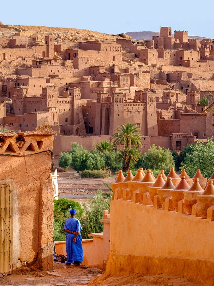 A person in a blue robe stands among orange walls, facing the ancient earthen fortress of Ait Ben Haddou with multiple towers and palm trees in a desert landscape—a scene straight from a travel guide inviting you to visit.