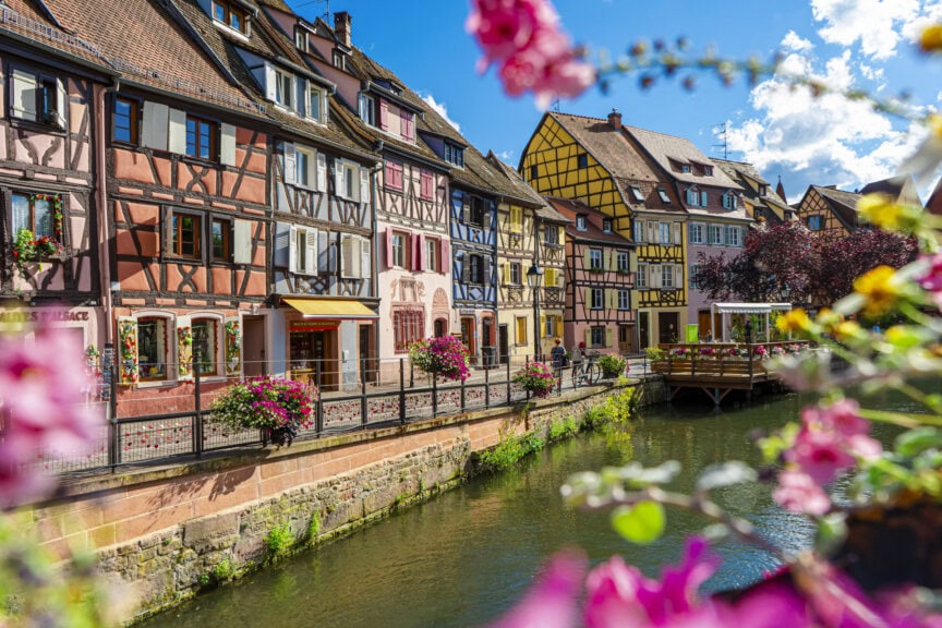 Colorful half-timbered houses line a canal under a bright sky, with flowers in the foreground and a wooden bridge on the right—a picturesque France travel scene perfect for visitors seeking the best time to visit this charming destination.