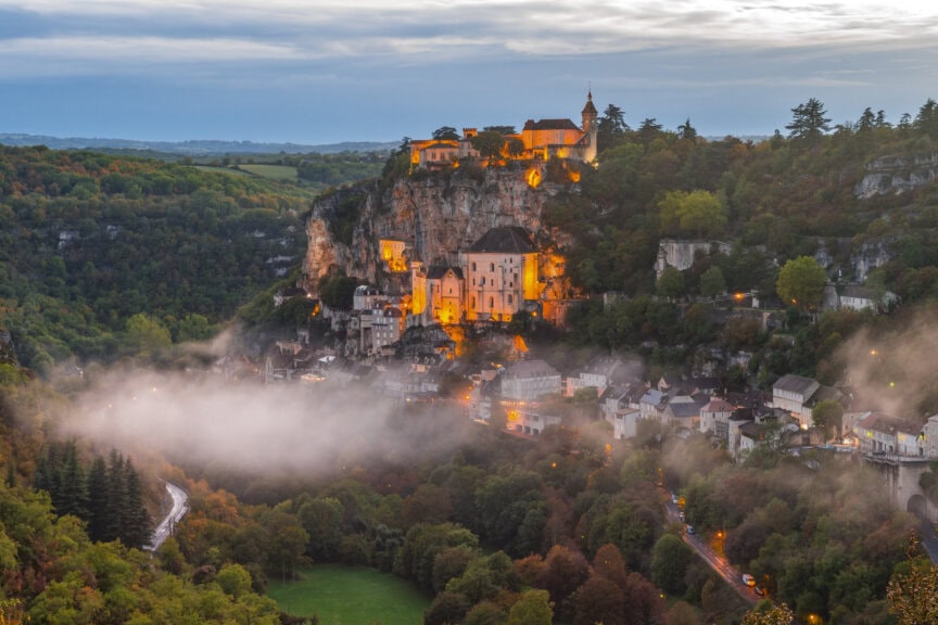 Hilltop village in France illuminated at dusk with mist rising below, surrounded by dense forest and a winding road in a valley—a perfect travel destination to explore during the best time to visit.
