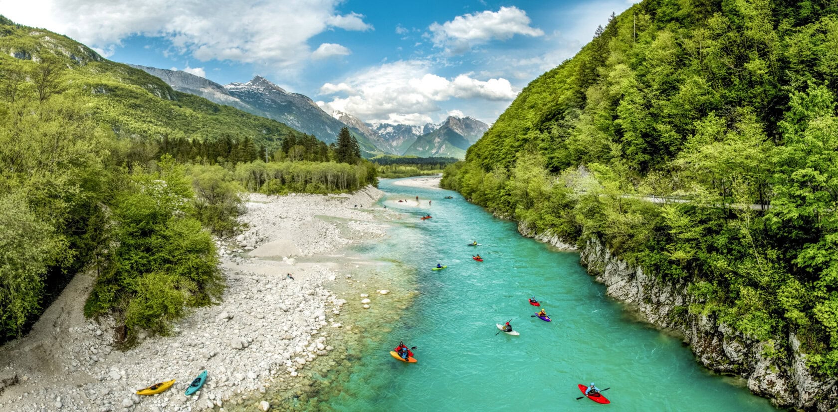 A group of kayakers paddles down a turquoise river, surrounded by green forests and mountains under a partly cloudy sky—showcasing the stunning scenery that makes spring or summer the best time to visit Slovenia.