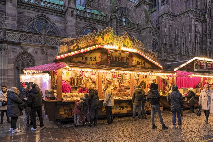 People stand in front of a brightly lit "Hansel & Gretel" market stall selling sweets at an outdoor Christmas market, with a gothic building in the background—a magical scene for France travel during the best time to visit.