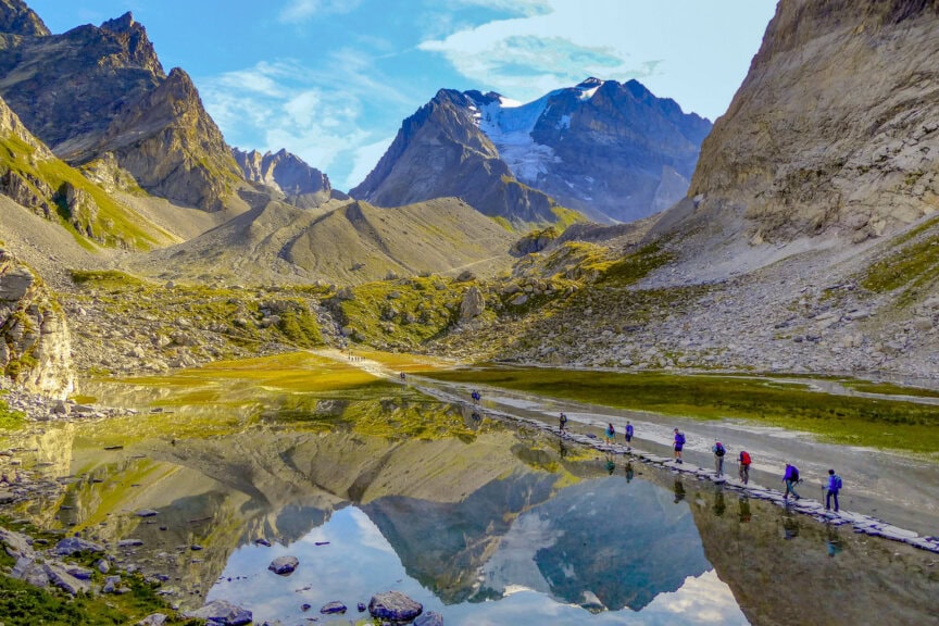 A group of hikers walks along a narrow path beside a calm mountain lake in France, with rocky peaks and blue sky reflected in the water—a scene best enjoyed during the best time to visit.