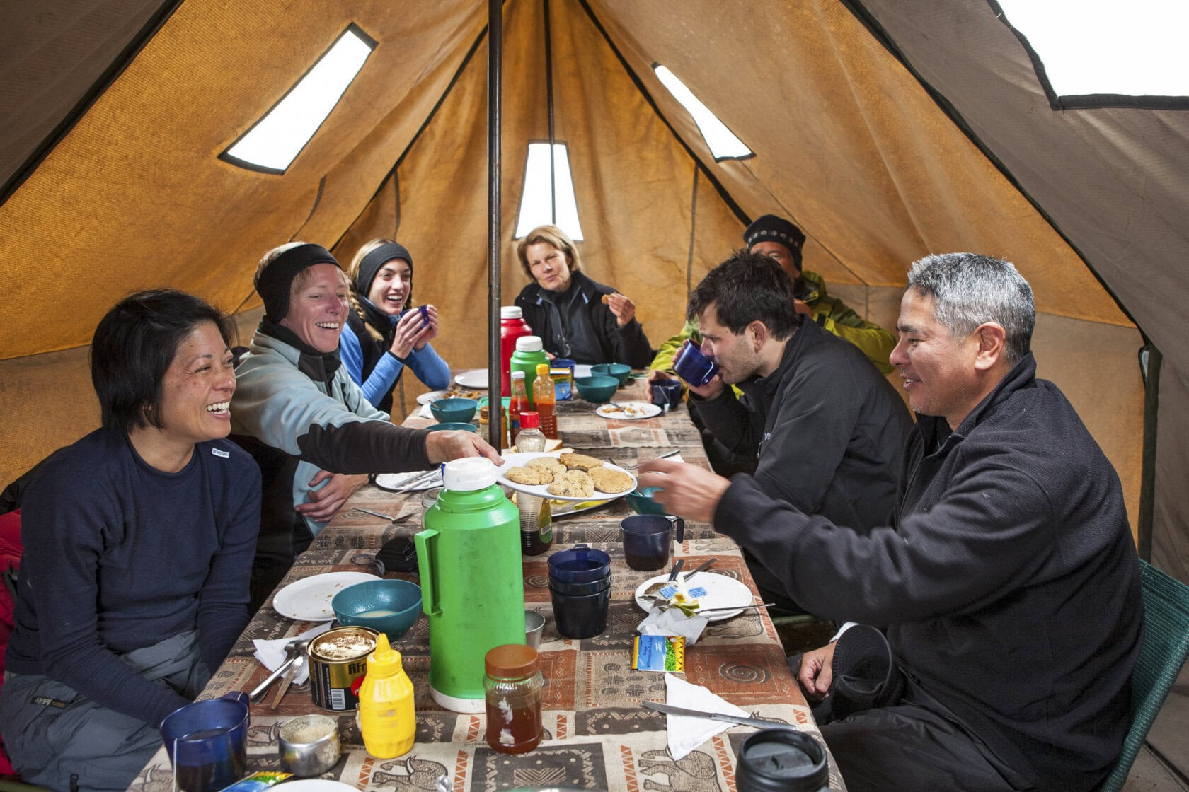 Six people sit around a table inside a tent, sharing a meal and smiling after a day of adventure on one of the renowned Kilimanjaro tours. The table is set with cups, plates, condiments, and food as the group appears to be thoroughly enjoying themselves.