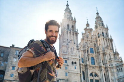 Man with a backpack smiles in front of an ornate cathedral with two tall towers on a sunny pilgrimage along the Camino de Santiago.