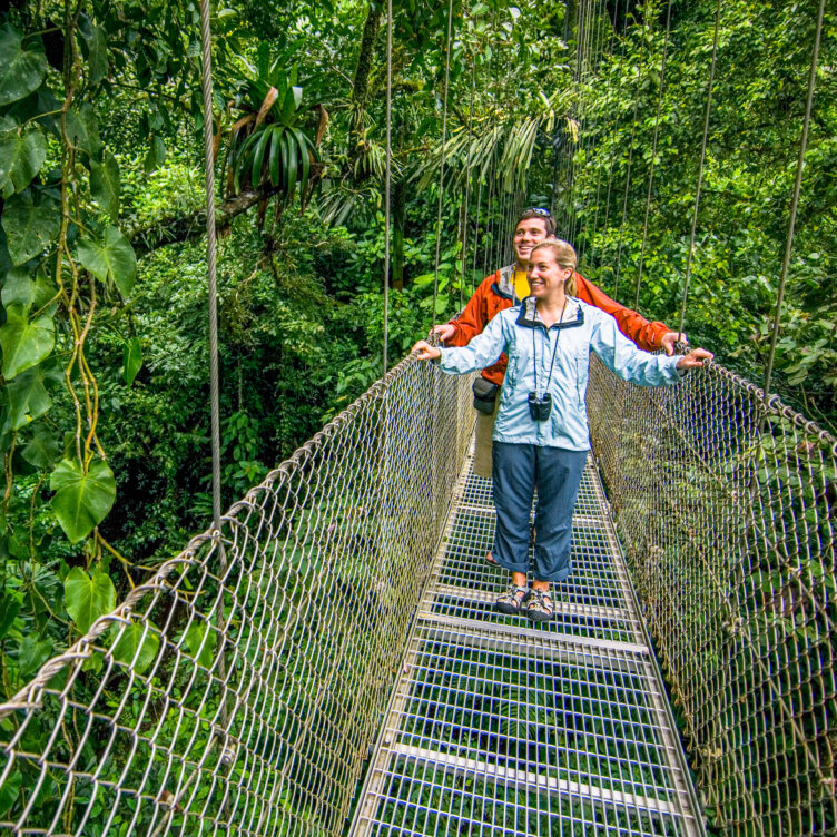 Two people walk on a suspension bridge surrounded by dense green foliage in Costa Rica, with one person holding binoculars and both looking ahead and smiling—an unforgettable moment for any traveler seeking valuable visitor information and travel tips before exploring the rainforest.