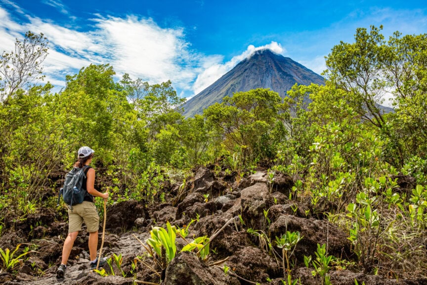 A person with a backpack hikes on a rocky trail surrounded by green vegetation, approaching a large, cloud-topped volcanic mountain under a blue sky—an unforgettable scene that captures the adventure awaiting in Costa Rica, especially during the best time to visit.