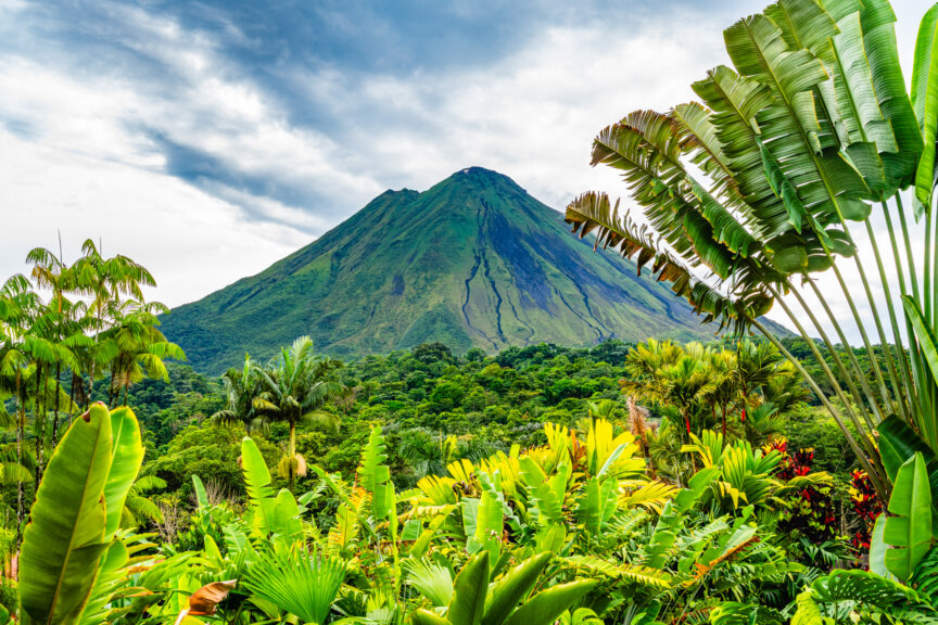 A volcanic mountain rises above dense green tropical vegetation under a partly cloudy sky, showcasing the natural beauty found in Costa Rica’s renowned parks and reserves.
