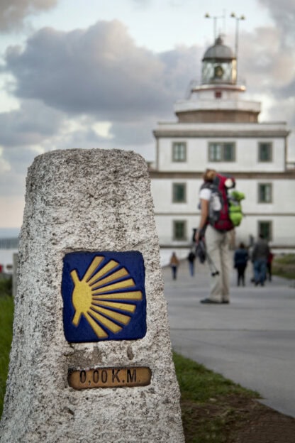 A stone marker with a yellow scallop shell symbol and “0,00 KM” marks the end of the Camino de Santiago pilgrimage; a person with a backpack walks in the background.