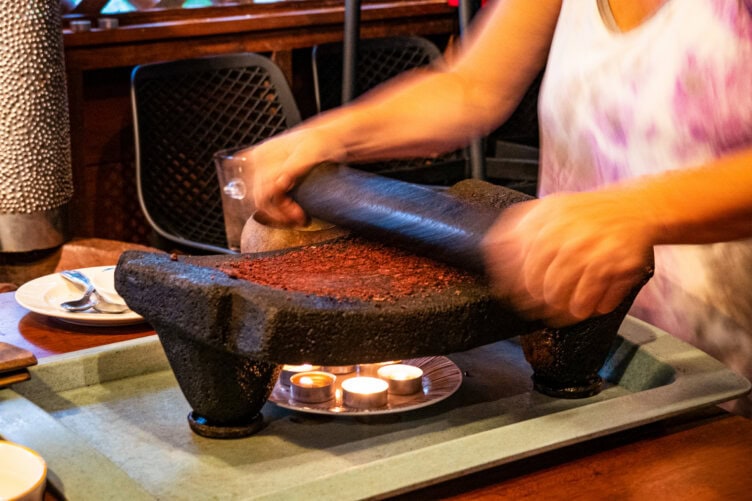 A person uses a stone grinder to crush cocoa beans on a table, evoking the traditional flavors enjoyed during Costa Rica travel, with small candles and plates nearby.