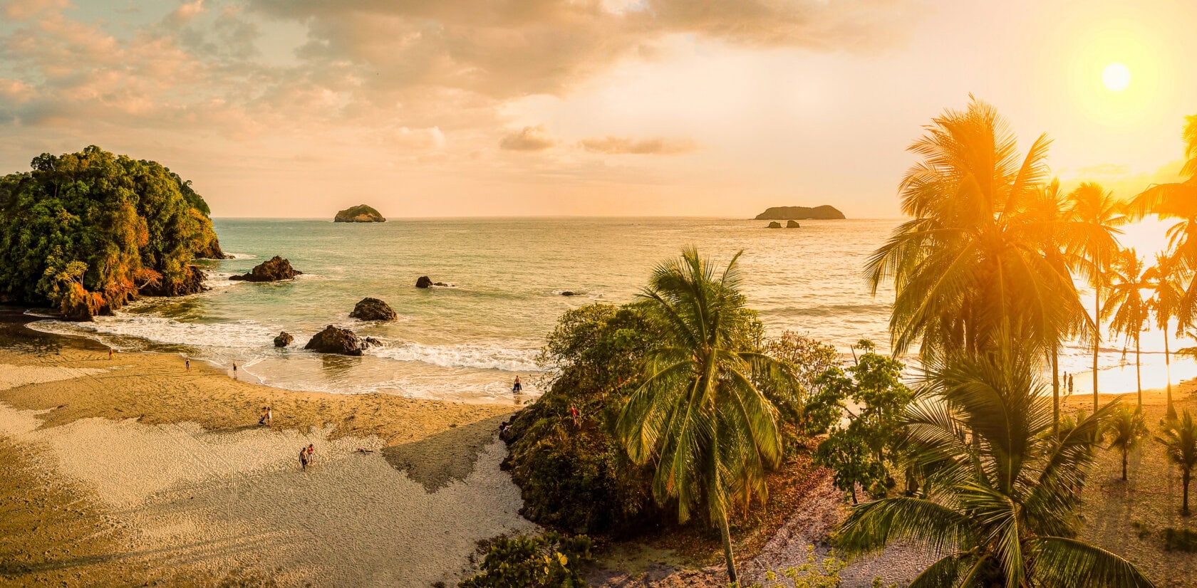 Sunset over a tropical Costa Rica beach with palm trees, small islands in the distance, gentle waves, and a few people walking on the sand. Experience this serene scene during the best time to visit for perfect weather and breathtaking views.