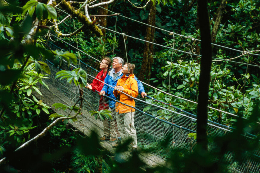 Three people in colorful jackets stand on a suspension bridge in a dense, green forest, looking up at the canopy—a scene reminiscent of the lush reserves and parks found throughout Costa Rica.