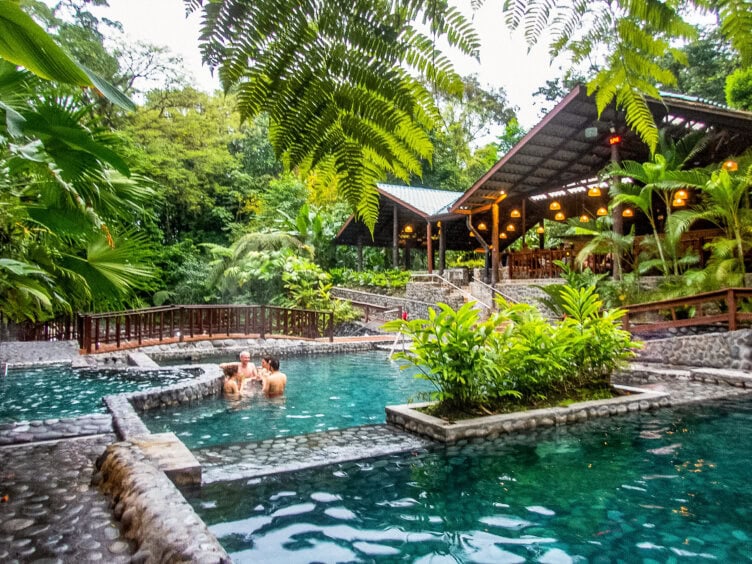 People relax in a stone-lined pool surrounded by lush tropical plants, with a covered pavilion and wooden walkway nearby—a perfect snapshot of travel in Costa Rica. This inviting spot is one of the top things to do for visitors seeking tranquility and natural beauty.