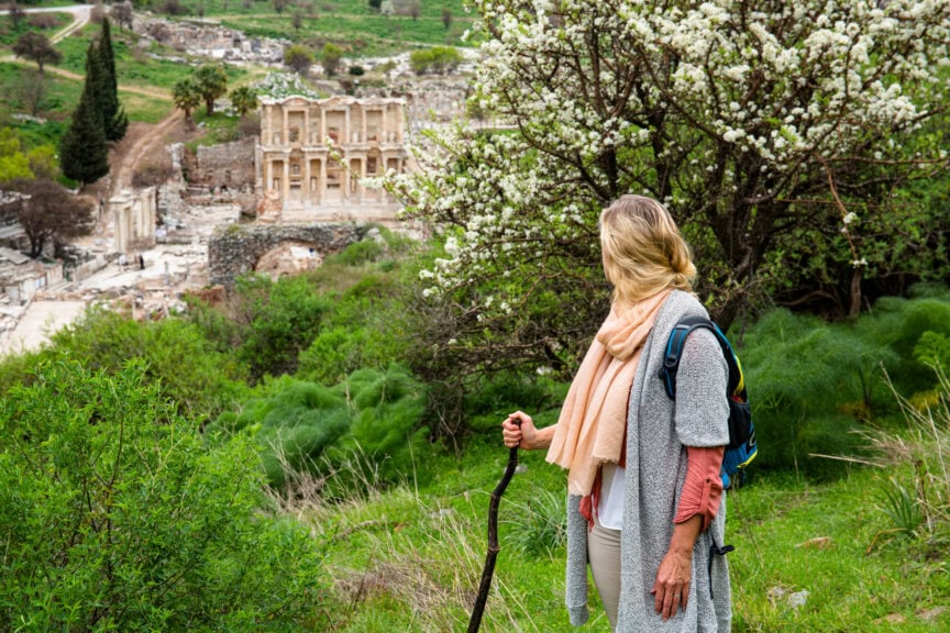 A person with a walking stick stands on a grassy hillside, looking at ancient ruins and blooming trees in the background—capturing the beauty that awaits travelers during the best time to visit Turkey.