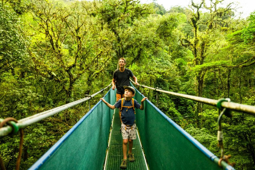 An adult and a child walk on a suspended bridge through dense green forest in Costa Rica, looking up at the trees around them and enjoying the vibrant jungle scenery—the best time to visit is when the rainforest is at its lushest.