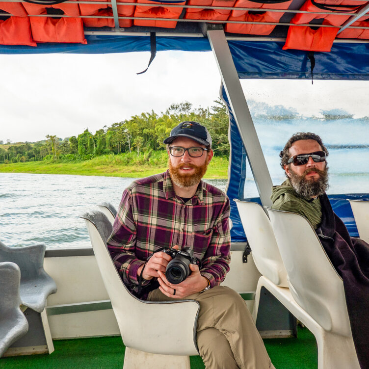 Two men sit on a boat in Costa Rica; one holds a camera and wears glasses and a cap, while the other wears sunglasses. Green landscape and water are visible outside the boat—a picture-perfect moment that captures why travel tips and know before you go advice can make your journey unforgettable.