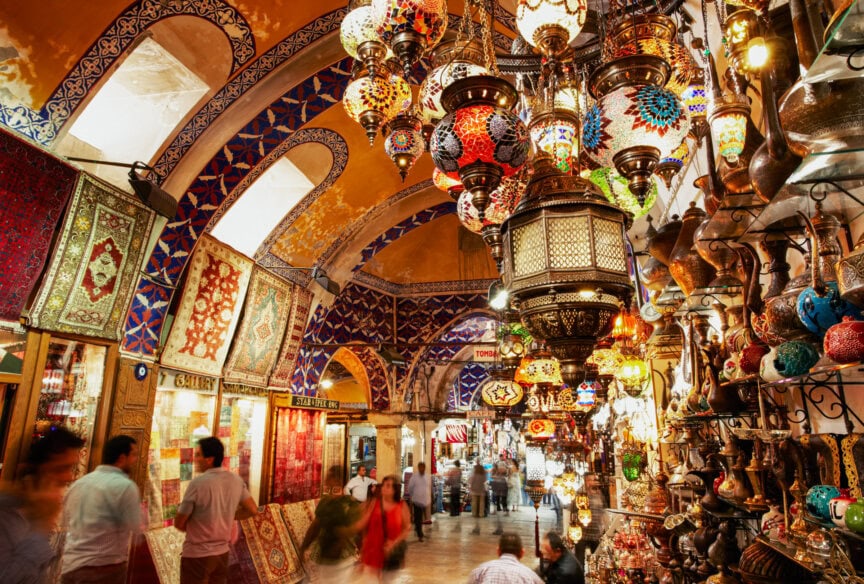 Interior view of a bustling market with colorful mosaic lamps, patterned rugs, and ornate decorations displayed under arched ceilings. Shoppers walk through the vibrant scene, capturing the lively atmosphere that makes exploring these markets one of the highlights during the best time to visit Turkey.