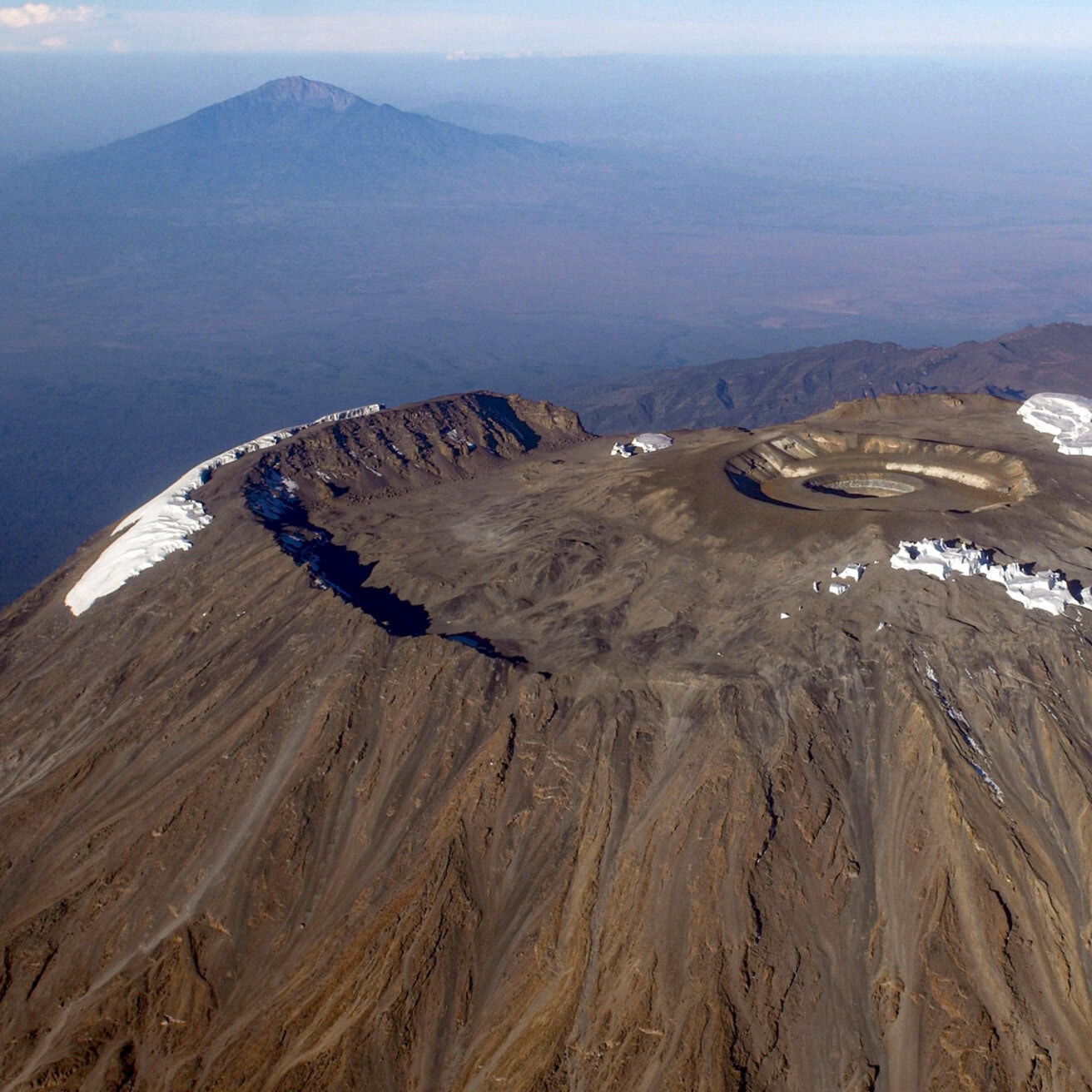 Aerial view of Mount Kilimanjaro’s volcanic crater with patches of snow, distant mountain and flat plains visible in the background—an iconic scene that hints at the once-in-a-lifetime experience of crater sleeping high above the African landscape.